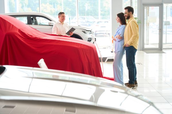 Car dealership consultant presents a car under a red cover, a buyer and his wife are waiting for a surprise