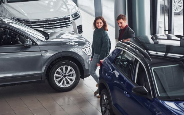 Professional salesman assisting young girl by choosing new modern automobile indoors.