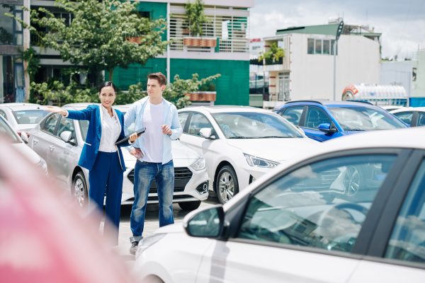 Pretty smiling young Asian manager walking with customer among rows of cars and telling about advantage and disadvantages of each one